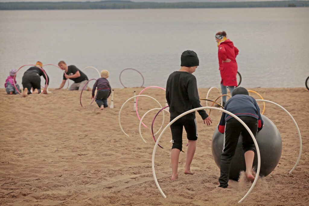 Lapset ja aikuiset pyörittävät suurta palloa renkaiden läpi Ärjän rantahietikolla.